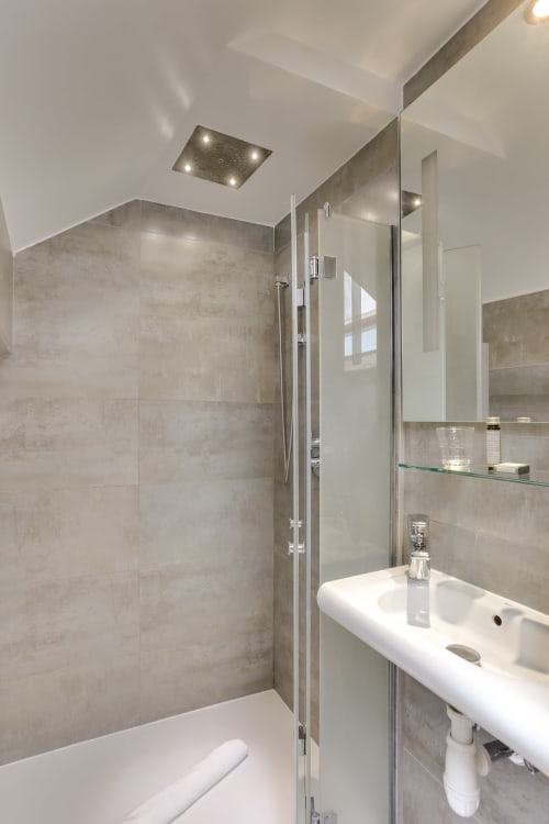 Salle de bains moderne à l'Hôtel Louvre Richelieu Paris avec douche sans cadre, lavabo blanc et finitions carrelage neutre