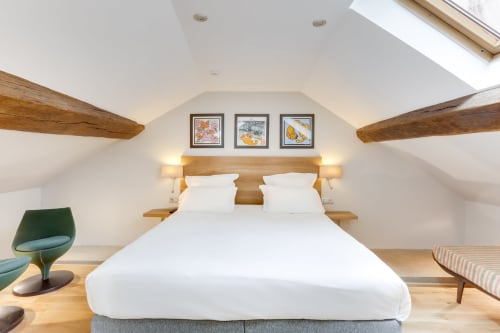 Chambre mansardée lumineuse à l'Hôtel Louvre Richelieu Paris avec poutres apparentes, verrière, lit queen-size et mobilier moderne.