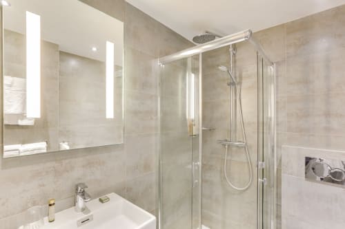 Salle de bain moderne de l'Hôtel Louvre Richelieu Paris avec douche ouverte, miroir lumineux et lavabo blanc