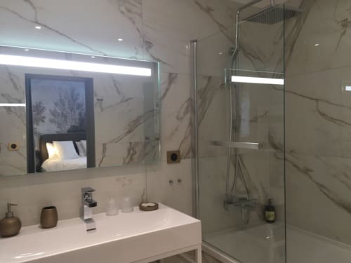 Modern bathroom at Le château d'Hermonville with marble walls, glass shower, lit mirror, and view of bedroom.