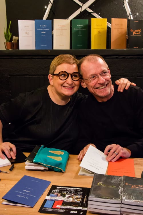 Deux personnes souriantes lors d'une séance de signature de livres, avec des ouvrages colorés exposés sur une étagère derrière elles aux Filles du Bord de Mer