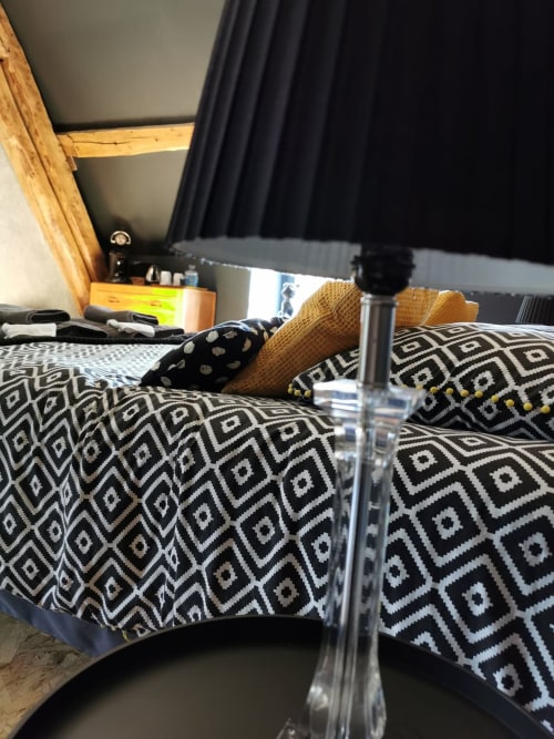 Chambre confortable à La Cour d'Hortense avec literie à motifs, poutres en bois, carafe d'eau et rideaux noirs