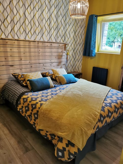 Chambre moderne de La Cour d'Hortense aux murs moutarde, literie géométrique, tête de lit en bois et vue sur le jardin