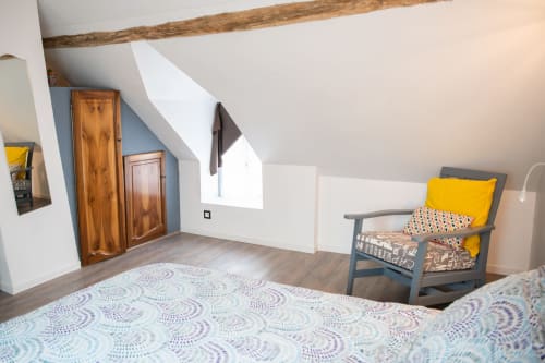 Chambre cosy sous les combles aux Gîtes des Châteaux avec parure à motifs, poutres bois, fauteuil gris et portes de penderie en bois