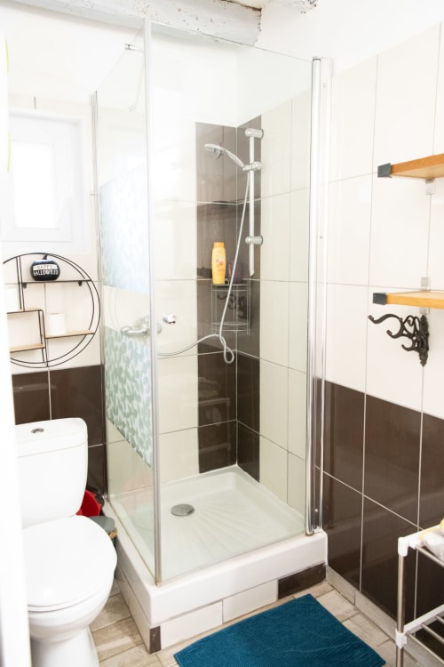 Salle de bain moderne avec douche vitrée, carrelage foncé, toilettes blanches et étagères en bois aux Gîtes des Châteaux