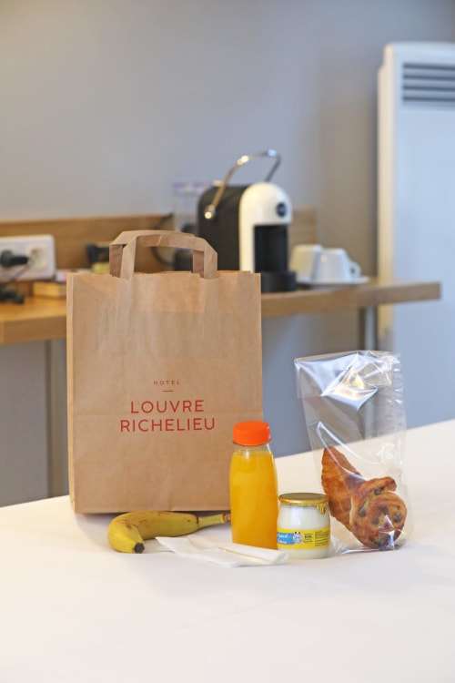Petit-déjeuner bienvenue à l'Hôtel Louvre Richelieu : viennoiseries, jus, banane et sac papier aux couleurs de l'hôtel dans la chambre parisienne