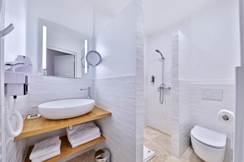 Salle de bains moderne de l'Hôtel Louvre Richelieu Paris avec carrelage blanc, lavabo rond, meuble vasque en bois et espace douche