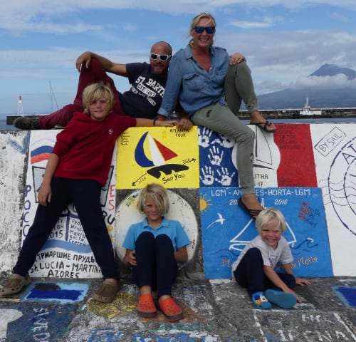 Family of six posing at decorated wall in Pleyber-Christ, France with mountain view and boats in background