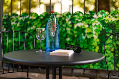 Table de patio extérieure chez Un Air De Famille à Vittel avec bouteille d'eau, verre de vin et lunettes de soleil surplombant un mur de lierre verdoyant