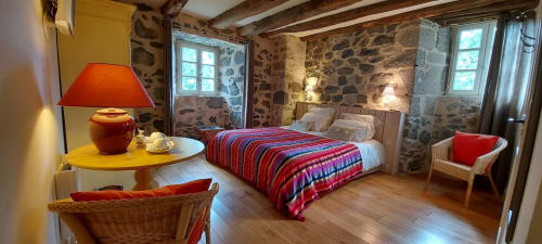 Chambre spacieuse au Mas De Rigoulac avec murs en pierre, literie rayée rouge, poutres bois et vue jardin en Aubrac