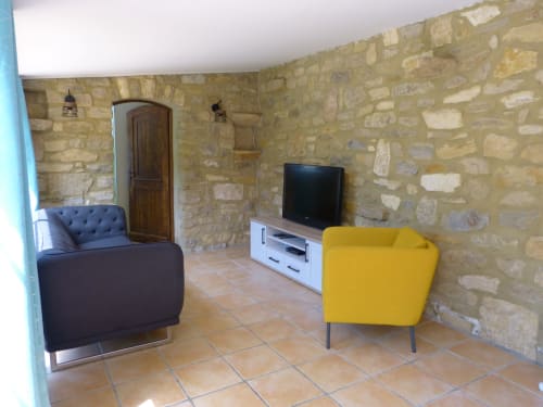 Stone-walled living room at Villa Thébaïde with blue and yellow armchairs, TV, and terracotta tile flooring in Laudun