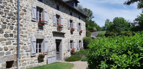 Façade en pierre du gîte Le Mas De Rigoulac à Argences-en-Aubrac avec jardinières fleuries rouges, volets blancs et jardins verdoyants