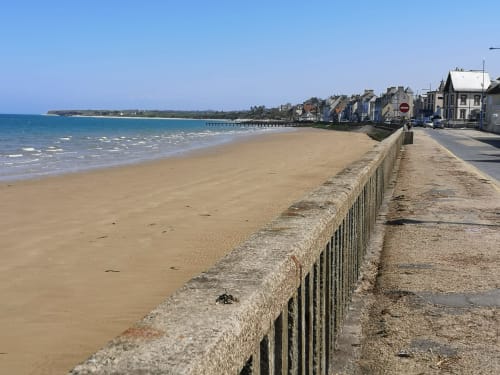 Sandy beach at Isigny-sur-Mer with stone wall, sea, and waterfront buildings under clear blue sky