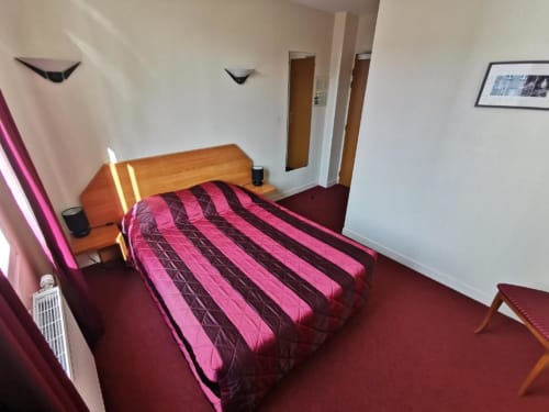 Habitación de hotel moderno en La Consigne en Caen con cama de madera, ropa de cama de rayas rosas, alfombra burdeos e iluminación de neón.