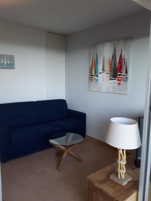 Coin salon cosy dans l'appartement d'Ajaccio avec canapé bleu marine, tableau de voilier et lampadaire blanc avec vue sur les Sanguinaires de Corse
