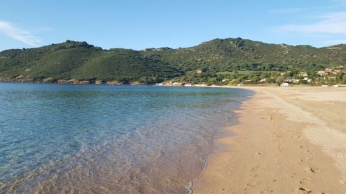 Plage de sable en Corse avec une eau cristalline, des collines verdoyantes et des maisons dispersées le long du littoral