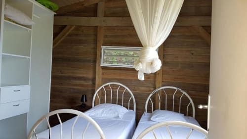 Dos camas gemelas con marcos metálicos blancos en un bungalow de madera en Le Bougainvillier Blanc, Pointe Noire, con cortinas translúcidas y vistas al mar