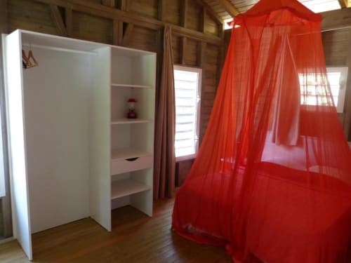 Cottage bedroom at Le Bougainvillier Blanc with white wardrobe, red mosquito canopy bed, and wooden beams in Pointe Noire