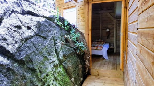 Entrada de cabaña de madera en Le Bougainvillier Blanc con muro de piedra cubierto de musgo y vista del interior del dormitorio, Pointe Noire