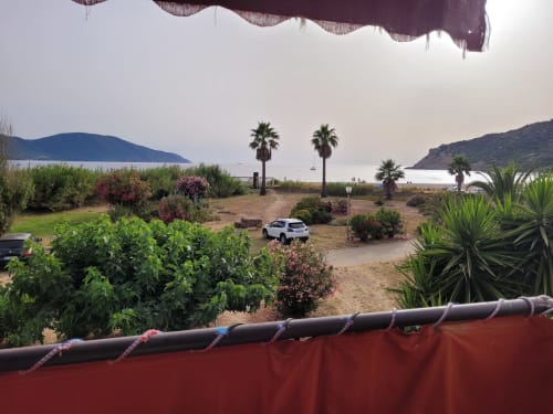 Jardin face mer d'une maison d'hôtes corse avec palmiers, voiture blanche, terrain de sable donnant sur la baie méditerranéenne