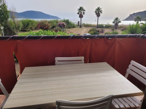 Terrasse de maison d'hôtes corse avec table en bois, chaises blanches, paravent rouge, vue sur mer et palmiers