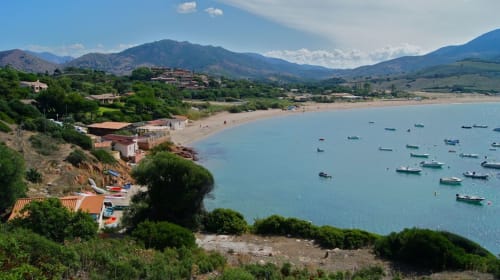 Baie côtière corse avec bateaux colorés, plage de sable, villas sur les collines et montagnes sous ciel bleu