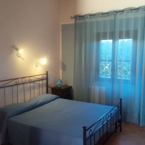 Camera luminosa a Podere Oca Bianca con letto in metallo, tende blu trasparenti, vista sul giardino e applique murali