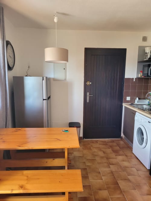 Cuisine d'appartement corse lumineuse avec table à manger en bois, réfrigérateur inox, machine à laver et porte en bois foncé