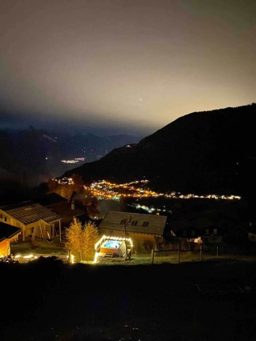 L'auberge La Tzoumaz la nuit, chalet illuminé, bain à remous et lueurs du village dans la vallée alpine suisse