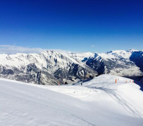 Piste de ski enneigée à La Tzoumaz avec pics alpins et balises oranges sous un ciel bleu dégagé