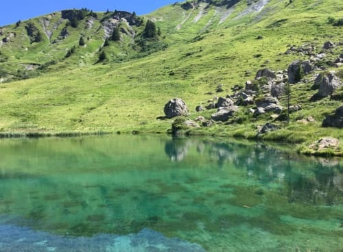 Crystal-clear alpine lake with lush green hillside, rocks, and mountain slopes at La Maison De L'Épine, Aime la Plagne