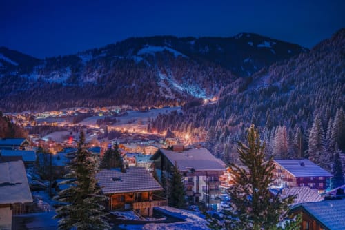 Chalet Domisiladoré nestled in snowy Alpine mountains at dusk, surrounded by illuminated chalets and frosted forest near