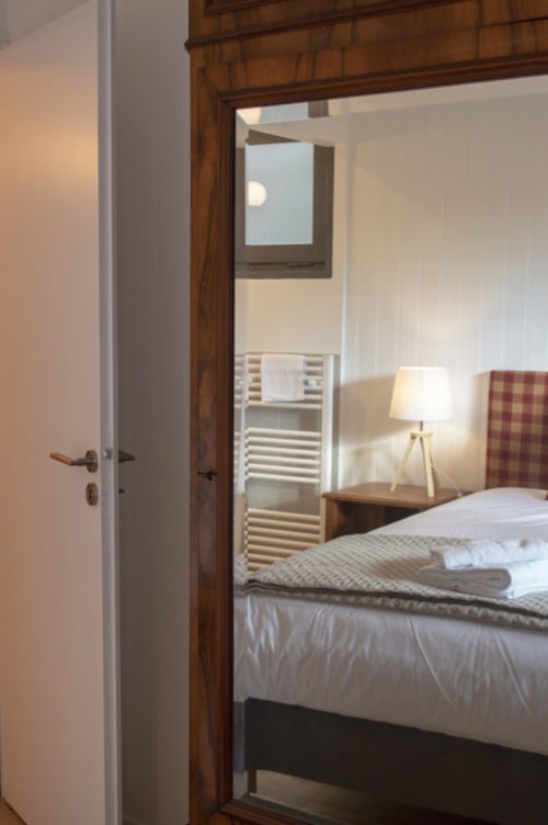 Bedroom entrance at Chalet Domisiladoré showing bed with plaid headboard, bedside lamp, and bathroom with towel rack