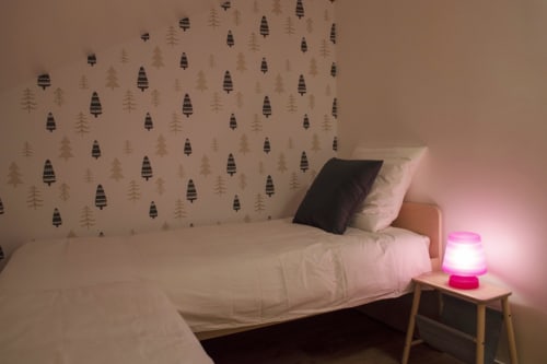 Chambre chaleureuse au Chalet Domisiladoré avec papier peint forêt, literie blanche, éclairage rose et table de nuit en bois.