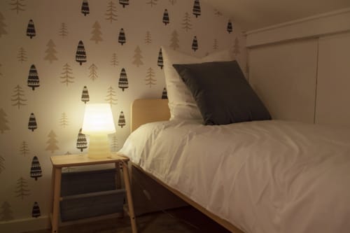 Chambre cosy du Chalet Domisiladoré avec papier peint forêt, literie blanche, oreiller noir, table de nuit en bois et lampe chaleureuse