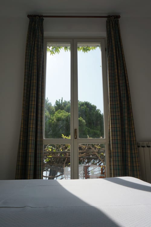 Helles Schlafzimmer mit karierten Vorhängen, die Glastüren mit Blick auf grüne Bäume und Meerblick im Hotel Beau Rivage in Alassio rahmen