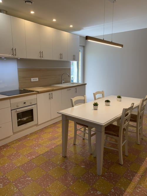 Modern cottage kitchen with white cabinetry, wooden backsplash, dining table with chairs, and stone floor at Gites Mas D'en
