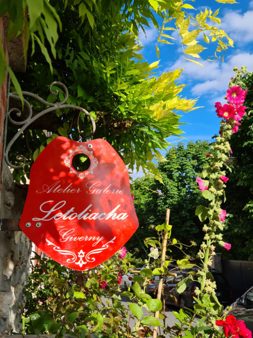 Enseigne rouge de l'Atelier Galerie Lelobacha à Giverny, entouré de fleurs colorées et de verdure luxuriante