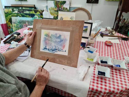 Artiste peignant à l'aquarelle au Petit Nid d'Aigle, gîte créatif à Giverny, avec œuvres encadrées et fournitures artistiques sur la table.