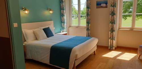 Chambre lumineuse au Château de Grandmont avec mur turquoise, lit blanc, chemin de lit bleu, appliques en laiton et vue sur le jardin