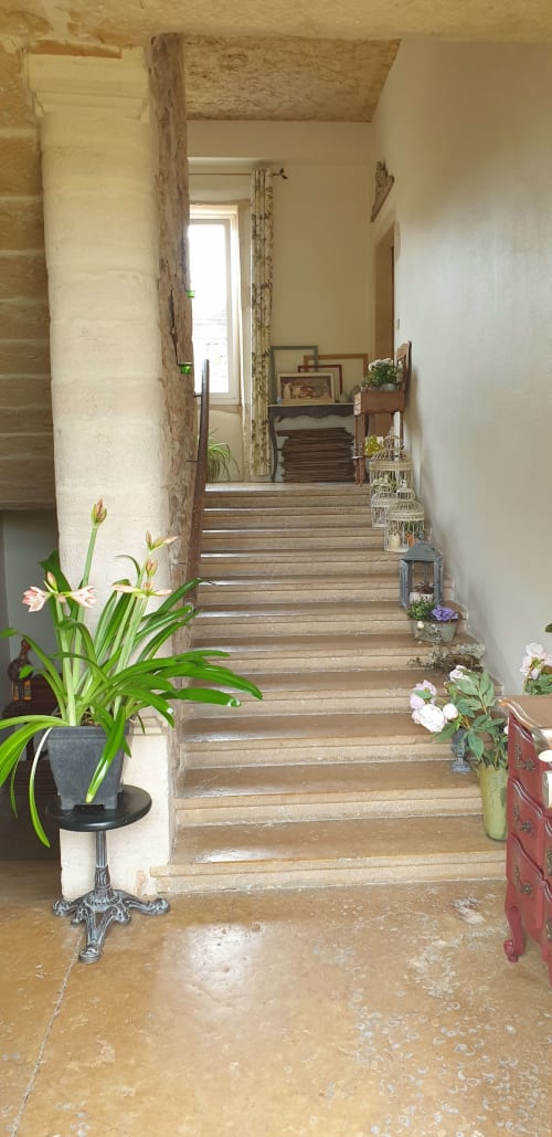 Couloir en pierre brute avec escalier en bois, fleurs en pots et lumière naturelle au gîte Château de Grandmont à Blacé