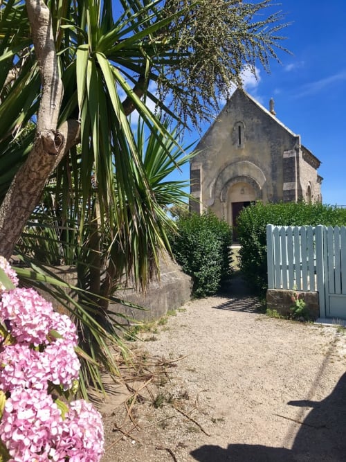 Entrée de la maison d'hôtes L'Ermitage à Saint-Vaast-la-Hougue avec chapelle en pierre, fleurs roses et palmiers
