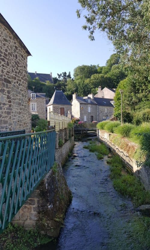 Bâtiments en pierre au bord du ruisseau aux Forges Du Lou B&B à Jugon-les-Lacs, balustrade verte et arbres luxuriants