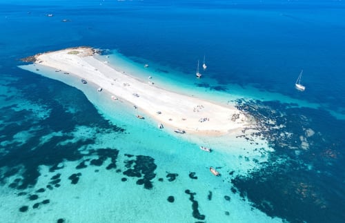 Aerial view of white sandy beach with turquoise waters, sailboats, and rocky outcrops near La Fôret-Fouesnant