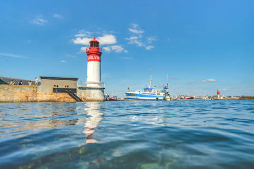 Red and white lighthouse at La Fôret-Fouesnant harbor with blue fishing boat and clear sky