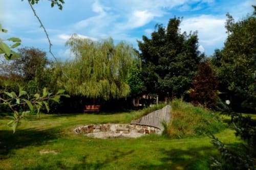 Lush garden at Aux Prés Du Berry B&B in Chavin, France, featuring fire pit, wooden deck, and mature trees under blue sky