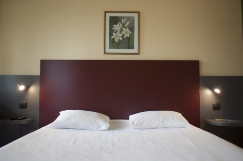 Chambre d'hôtel moderne à Mape Hotel avec tête de lit bourgogne, literie blanche, tables de nuit sombres et tableau floral encadré au-dessus du lit