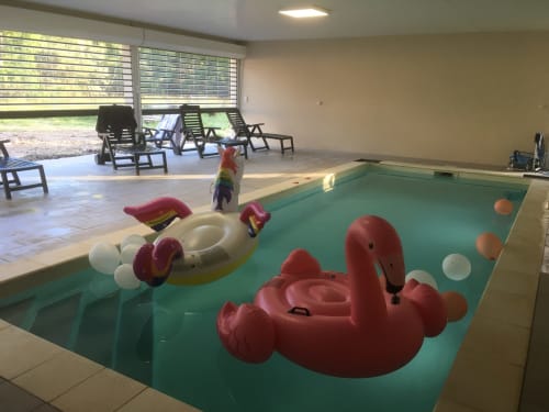 Piscine intérieure du Domaine Des Bois Argentés avec bouées colorées et transats près des fenêtres donnant sur le jardin