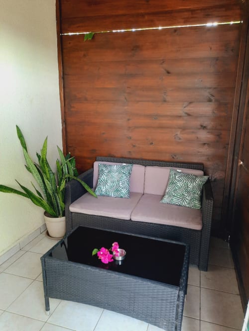 Cozy patio seating area at Gites Kaladja with dark wicker sofa, pink cushions, potted plant, and wooden backdrop in