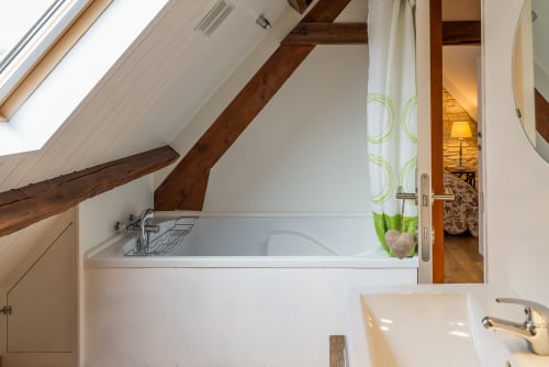 Salle de bain mansardée moderne avec baignoire balnéo, poutres apparentes et rideaux vue jardin au gîte Vue Sur Gindou, Gindou